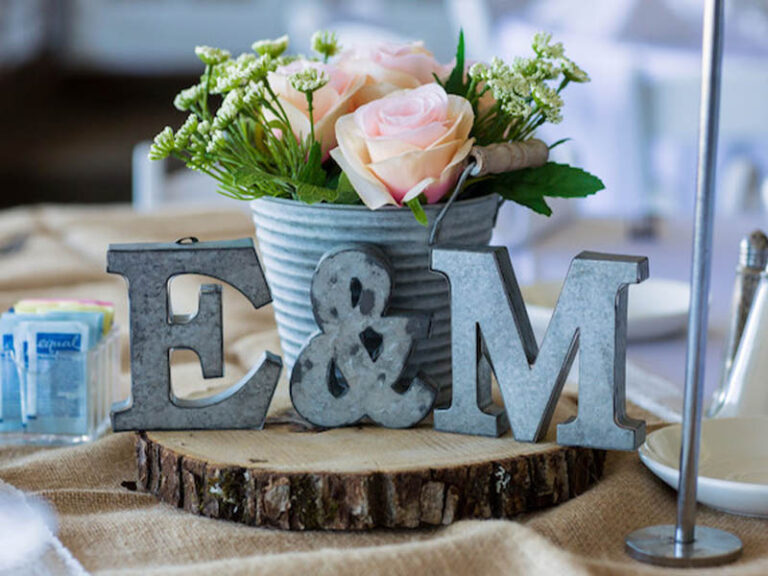 Rustic wedding centerpiece with pink roses, greenery, and metal initials on a wood slice.
